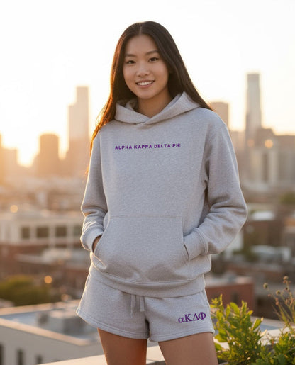 Person wearing light gray hoodie with purple ALPHA KAPPA DELTA PHI text and matching shorts outdoors