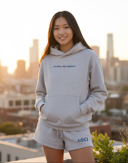 Person in gray Alpha Phi Omega hoodie and shorts on a city rooftop at sunset